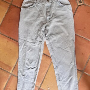 May include: Light blue denim jeans with a classic five-pocket design. The jeans have a button and zipper closure, and a straight leg fit. The pants are laid flat on a terracotta tile surface.