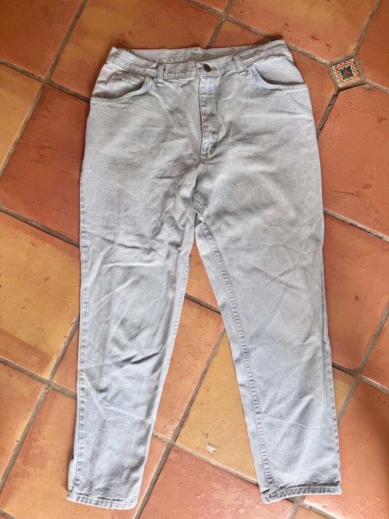 May include: Light blue denim jeans with a classic five-pocket design. The jeans have a button and zipper closure, and a straight leg fit. The pants are laid flat on a terracotta tile surface.