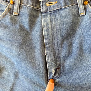 May include: Close-up of light blue denim jeans. The image shows the button closure, belt loops, and pockets. The jeans have a classic design with a worn appearance, and the button is a brass color.