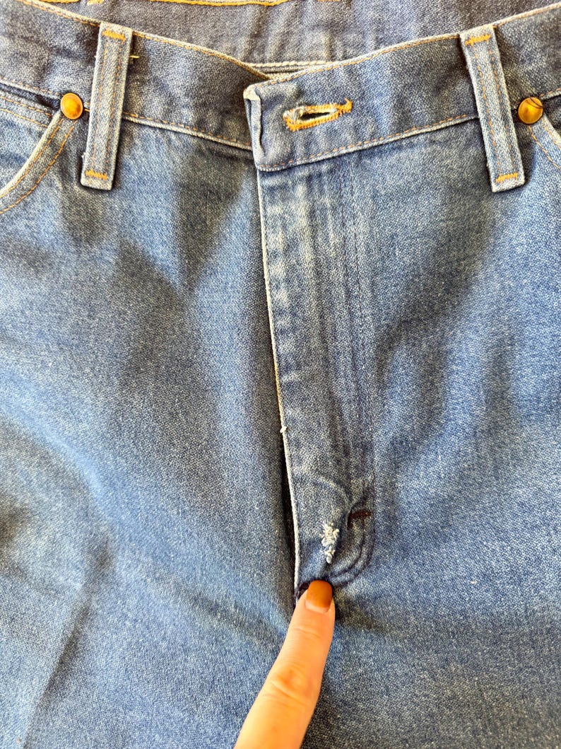 May include: Close-up of light blue denim jeans. The image shows the button closure, belt loops, and pockets. The jeans have a classic design with a worn appearance, and the button is a brass color.
