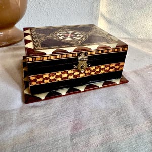 May include: A decorative wooden box with intricate inlaid patterns. The box features a geometric design on the lid and a contrasting pattern on the front. It has a small metal clasp and is set on a matching base. The box is a mix of dark brown, black, and cream colors.