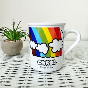 May include: White ceramic mug with a rainbow and clouds design. The mug has the name "CAROL" and the text "Song of Joy" printed on it.