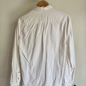 May include: A white long-sleeve shirt with a button-down collar, hanging on a wooden hanger. The shirt has buttoned cuffs and a classic design. The back of the shirt is visible, showing the yoke and pleats.