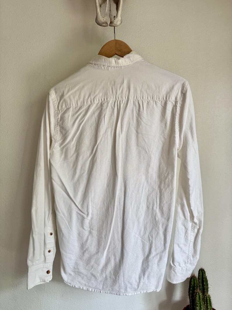 May include: A white long-sleeve shirt with a button-down collar, hanging on a wooden hanger. The shirt has buttoned cuffs and a classic design. The back of the shirt is visible, showing the yoke and pleats.