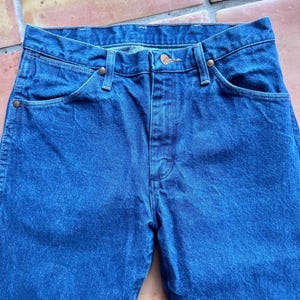 May include: Close-up of blue denim jeans. The jeans have a classic five-pocket design with copper-coloured rivets and a button closure. The fabric has a slightly faded appearance, suggesting wear.