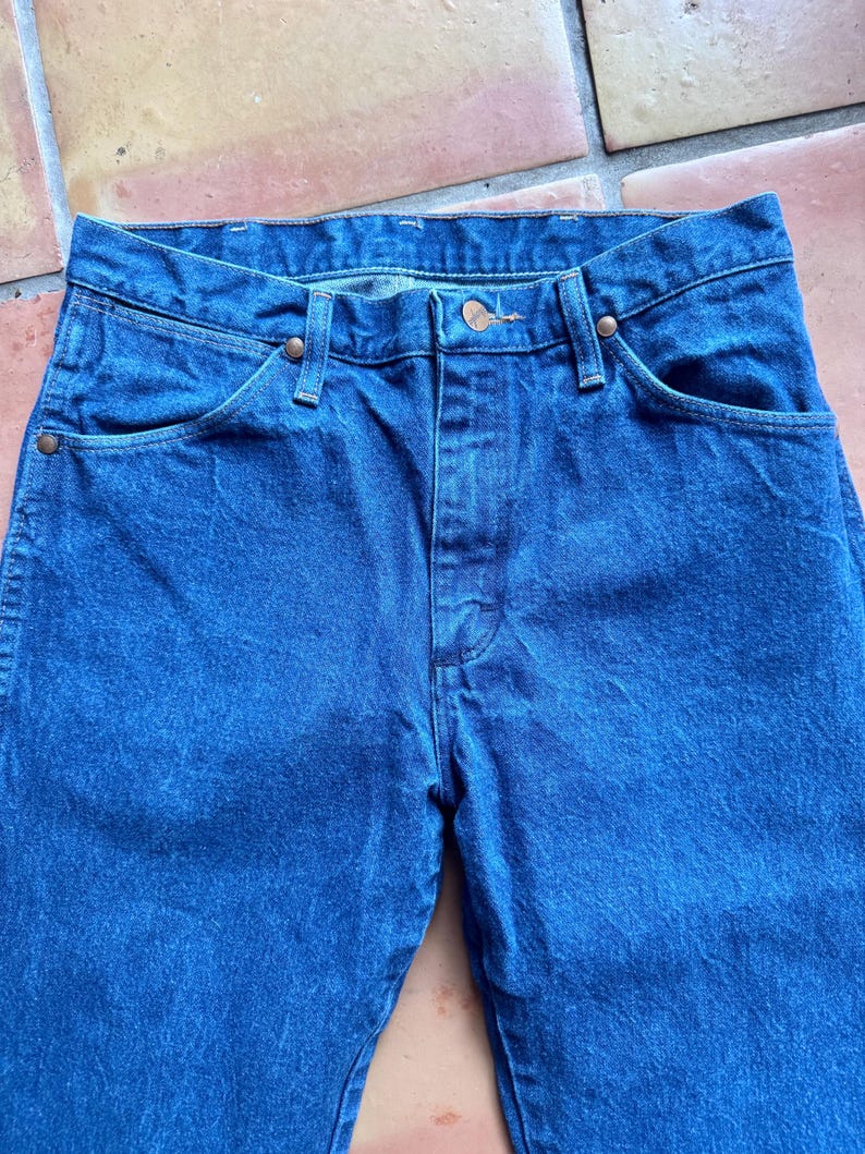 May include: Close-up of blue denim jeans. The jeans have a classic five-pocket design with copper-coloured rivets and a button closure. The fabric has a slightly faded appearance, suggesting wear.