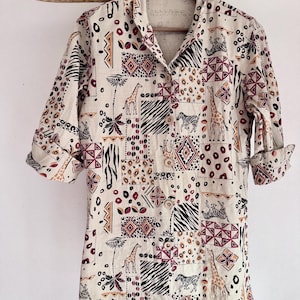 May include: A long-sleeve button-down shirt in a light beige fabric featuring a repeating patchwork pattern of giraffes, zebras, and abstract geometric shapes in black, dark red, and tan colors, with a pointed collar and cuffed sleeves.