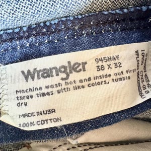 K&ouml;nnte beinhalten: Nahaufnahme eines Wrangler Jeansetiketts. Das wei&szlig;e Etikett zeigt den Markennamen, die Gr&ouml;&szlig;e 38 x 32 Zoll und Waschanweisungen. Die Jeans ist aus mittelblauem Denim mit sichtbaren N&auml;hten und einer Baumwollzusammensetzung.