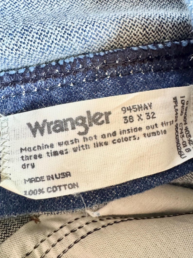 K&ouml;nnte beinhalten: Nahaufnahme eines Wrangler Jeansetiketts. Das wei&szlig;e Etikett zeigt den Markennamen, die Gr&ouml;&szlig;e 38 x 32 Zoll und Waschanweisungen. Die Jeans ist aus mittelblauem Denim mit sichtbaren N&auml;hten und einer Baumwollzusammensetzung.