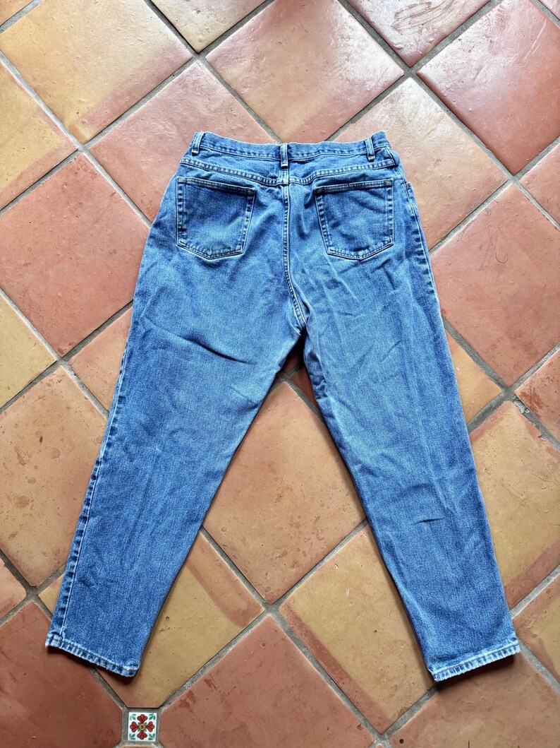 May include: Light blue denim jeans with a classic five-pocket design. The jeans are laid flat, showcasing the back pockets and the overall shape. The fabric has a slightly faded appearance, suggesting a vintage or worn look.