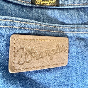 May include: Close-up of blue denim jeans with the Wrangler brand label. The label is a light brown rectangle with the Wrangler logo stitched in a cursive font. A smaller black label with the Wrangler logo is also visible.