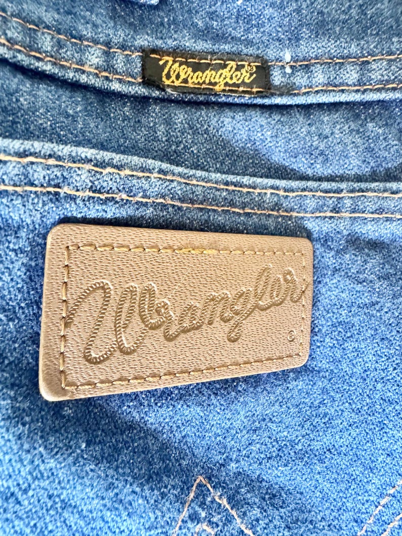 May include: Close-up of blue denim jeans with the Wrangler brand label. The label is a light brown rectangle with the Wrangler logo stitched in a cursive font. A smaller black label with the Wrangler logo is also visible.