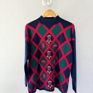 May include: A blue, green, and red patterned sweater with a mock neck and long sleeves. The sweater features a diamond pattern with paisley and floral embroidery.