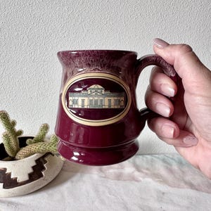 May include: Burgundy ceramic mug with a handle, featuring an oval design with a building illustration. The mug has a textured rim and a glossy finish. A small cactus in a decorative pot is visible in the background.