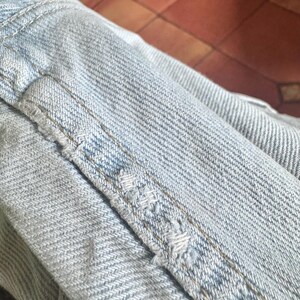May include: Close-up of light blue denim fabric with visible stitching and distressed details. The image highlights the texture and wear of the denim, showcasing the fabric's construction and the effects of use.