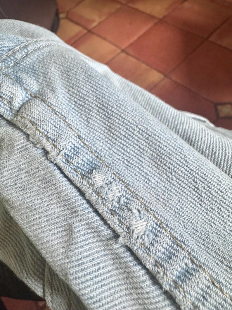 May include: Close-up of light blue denim fabric with visible stitching and distressed details. The image highlights the texture and wear of the denim, showcasing the fabric's construction and the effects of use.