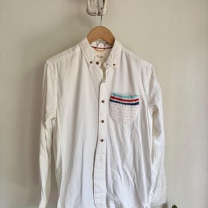 May include: A white button-down shirt with a crochet pocket featuring red, white, and blue stripes. The shirt has long sleeves and wooden buttons. The brand name "Koto" is visible on the collar.