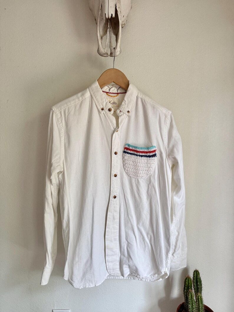 May include: A white button-down shirt with a crochet pocket featuring red, white, and blue stripes. The shirt has long sleeves and wooden buttons. The brand name "Koto" is visible on the collar.