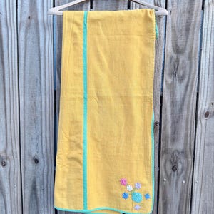 May include: A yellow textile with a turquoise border and a vertical stripe. The textile has a floral embroidery in shades of pink, blue, and white. The textile is hanging on a wooden hanger against a wooden fence.