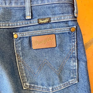 May include: Close-up of blue denim jeans with a Wrangler label on the back pocket. The jeans have a classic five-pocket design, with a brown leather patch featuring the Wrangler logo. The stitching is in a contrasting tan color.