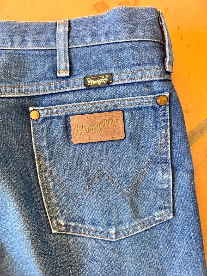 May include: Close-up of blue denim jeans with a Wrangler label on the back pocket. The jeans have a classic five-pocket design, with a brown leather patch featuring the Wrangler logo. The stitching is in a contrasting tan color.