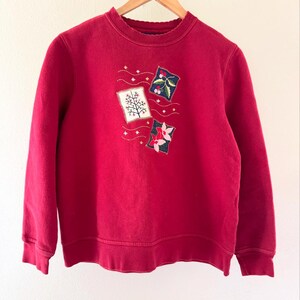 May include: A red sweatshirt with embroidered floral and tree designs. The sweatshirt has a crew neck and long sleeves.