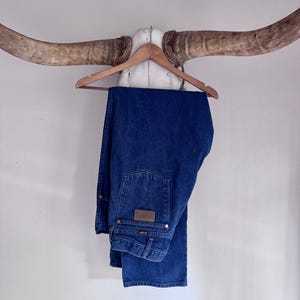 Puede incluir: Un par de pantalones vaqueros de mezclilla azul oscuro con un dise&ntilde;o cl&aacute;sico de cinco bolsillos, un parche de cuero marr&oacute;n en el bolsillo trasero y remaches met&aacute;licos. Los pantalones est&aacute;n doblados sobre una percha de madera.
