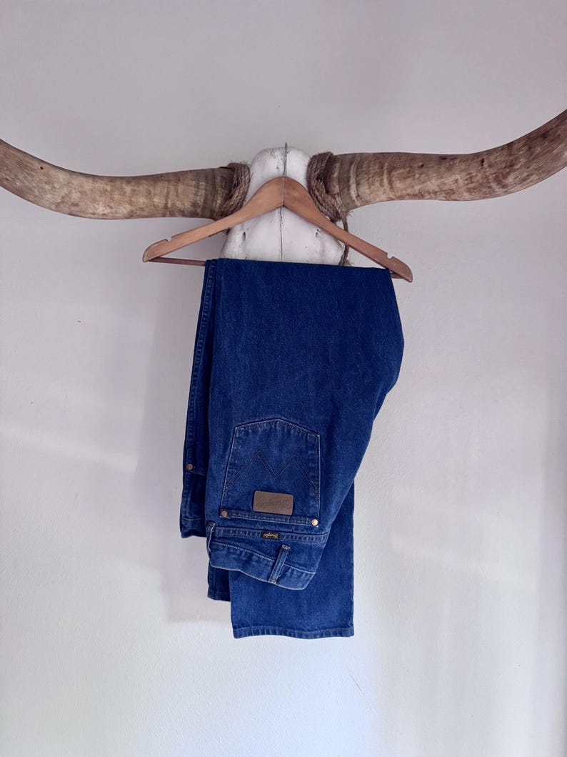Puede incluir: Un par de pantalones vaqueros de mezclilla azul oscuro con un dise&ntilde;o cl&aacute;sico de cinco bolsillos, un parche de cuero marr&oacute;n en el bolsillo trasero y remaches met&aacute;licos. Los pantalones est&aacute;n doblados sobre una percha de madera.