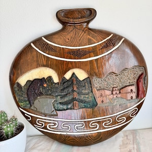May include: A wooden, oval-shaped wall hanging with a carved landscape scene. The dark brown wood features a handle and white accents. The landscape includes mountains, trees, and buildings, with a decorative border. The piece is likely a decorative item.