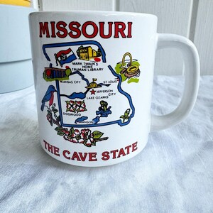 May include: White ceramic mug with a colorful illustration of the state of Missouri. The mug features the state's outline with various landmarks and cities labeled, including Kansas City, St. Louis, and Lake of the Ozarks. The text "Missouri" and "The Cave State" are printed on the mug.