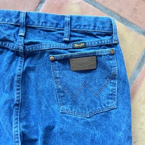 May include: Blue denim jeans featuring a high-waisted design with belt loops, a back pocket with a brown leather-like patch embossed with the Wrangler logo, copper-coloured rivets, and the signature W-shaped stitching on the back pocket.