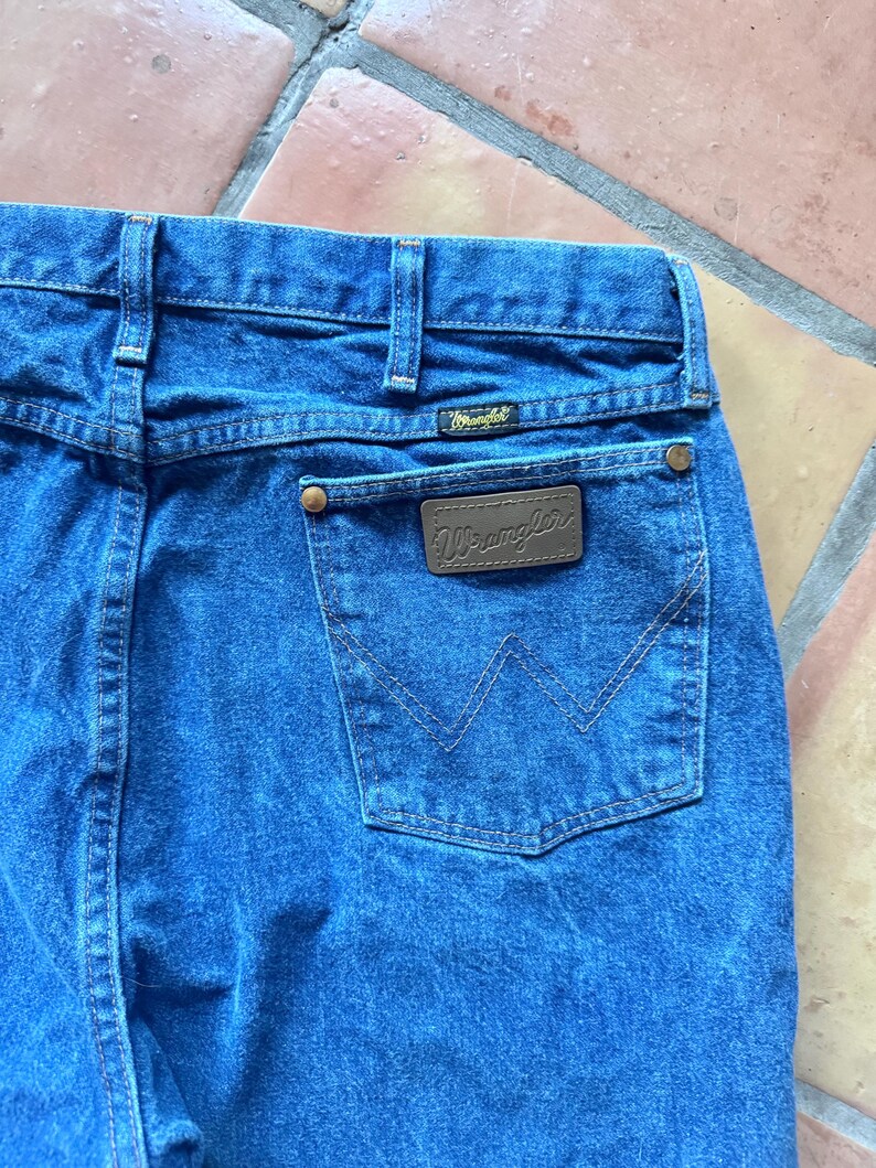 May include: Blue denim jeans featuring a high-waisted design with belt loops, a back pocket with a brown leather-like patch embossed with the Wrangler logo, copper-coloured rivets, and the signature W-shaped stitching on the back pocket.