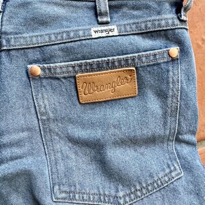 May include: Close-up of light blue denim jeans with the Wrangler brand label visible on the back pocket. The jeans have a classic five-pocket design, with copper-colored rivets and a leather patch. The Wrangler logo is also visible on the waistband.
