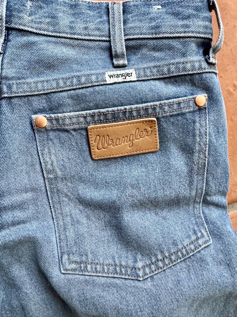 May include: Close-up of light blue denim jeans with the Wrangler brand label visible on the back pocket. The jeans have a classic five-pocket design, with copper-colored rivets and a leather patch. The Wrangler logo is also visible on the waistband.