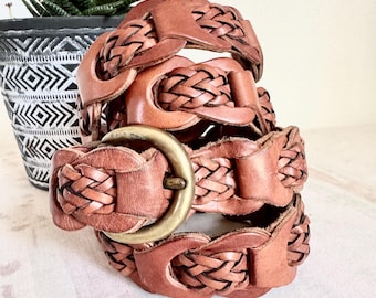 May include: A stack of three brown leather belts with a woven design. Each belt has a brass buckle. The belts are stacked on top of each other, showcasing the intricate braided pattern and the warm, earthy tones of the leather.