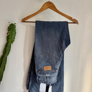 May include: Blue denim jeans hanging on a wooden hanger. The jeans have a classic five-pocket design, a brown leather patch with the brand name, and a straight leg fit. The jeans are displayed against a white wall with a cactus and a decorative element.