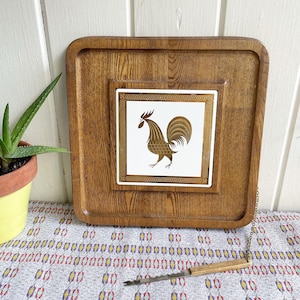 May include: A wooden serving tray with a ceramic tile featuring a gold rooster design. The tray has a chain attached to it and a small wooden handled cheese knife.
