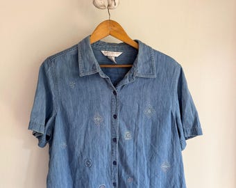 May include: A light blue denim shirt with short sleeves, embroidered details, and a button-down front. The shirt features geometric and floral embroidery in white and pink. The shirt is hanging on a wooden hanger.