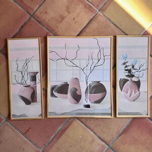 May include: A triptych of framed art prints featuring stylized vases in shades of pink and black. Each print has a gold frame and depicts vases with abstract designs and floral elements. The background is a soft pink with a window-like grid.