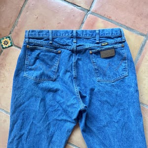 May include: A pair of blue denim jeans featuring a high-rise waistband, two back pockets with a signature W-shaped stitch pattern, copper-toned rivets, and a brown leather patch on the right pocket with the Wrangler brand name printed in gold.