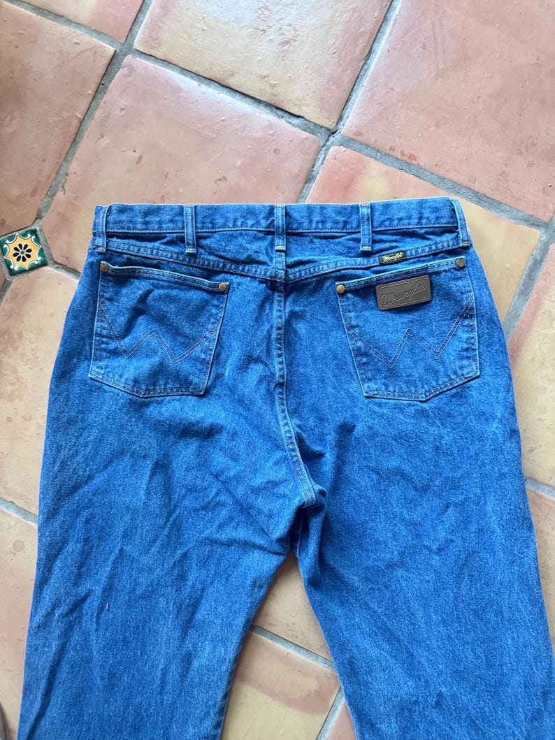 May include: A pair of blue denim jeans featuring a high-rise waistband, two back pockets with a signature W-shaped stitch pattern, copper-toned rivets, and a brown leather patch on the right pocket with the Wrangler brand name printed in gold.