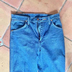 May include: Close-up of blue denim jeans with a button and zipper closure. The jeans have front pockets and copper-colored rivets. The fabric is a medium wash denim.