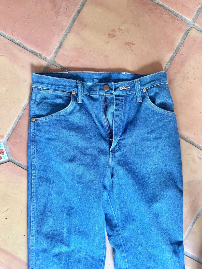 May include: Close-up of blue denim jeans with a button and zipper closure. The jeans have front pockets and copper-colored rivets. The fabric is a medium wash denim.