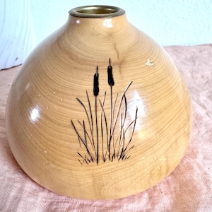 May include: A rounded wooden vase with a smooth, light-toned finish and a metallic rim at the opening. The exterior features a dark, etched illustration of two cattails surrounded by tall, thin blades of grass, showcasing natural wood grain.