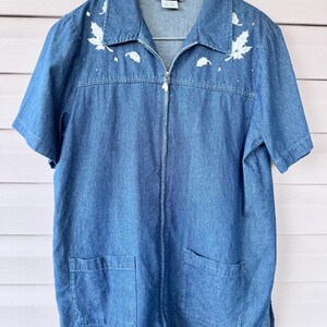 May include: A medium-wash blue denim shirt with a full-length front zipper, short sleeves, and two lower front pockets. The upper chest features white embroidered maple leaf motifs and small silver-toned studs. The label reads JASON SMITH SPORT.