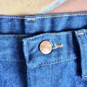 K&ouml;nnte beinhalten: Nahaufnahme einer blauen Jeans mit Knopfverschluss. Der Knopf ist hellbraun und tr&auml;gt die Gravur "Wrangler". Die Jeans hat sichtbare N&auml;hte.