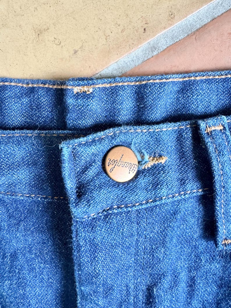 K&ouml;nnte beinhalten: Nahaufnahme einer blauen Jeans mit Knopfverschluss. Der Knopf ist hellbraun und tr&auml;gt die Gravur "Wrangler". Die Jeans hat sichtbare N&auml;hte.