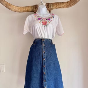 May include: A white embroidered top with a colorful floral design and a denim button-down skirt. The skirt is a medium wash blue and has a classic A-line shape. The outfit is displayed on a mannequin.