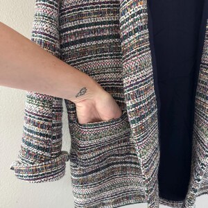 May include: A multi-colored tweed blazer with a navy blue lining. The blazer features a textured weave with hues of green, purple, and white. The sleeve cuff is turned up, and a hand is in the pocket.