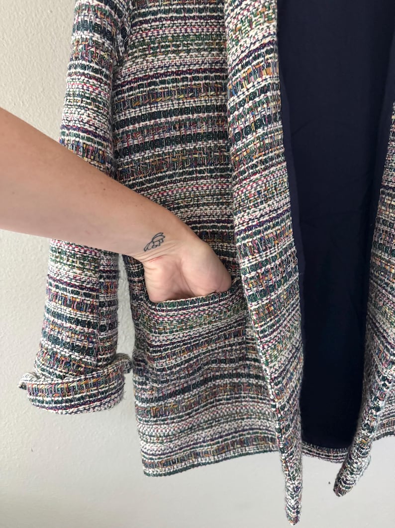 May include: A multi-colored tweed blazer with a navy blue lining. The blazer features a textured weave with hues of green, purple, and white. The sleeve cuff is turned up, and a hand is in the pocket.